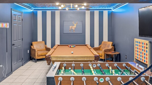 Een kamer met een pooltafel, twee fauteuils, een tafelvoetbalspel, een lijst met een foto van een hert en een televisie aan de muur.