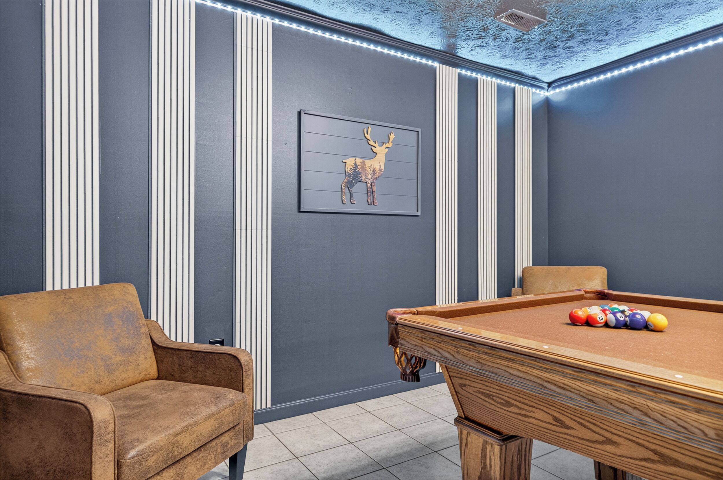 A pool table with balls, a sofa, and a framed deer picture in a room with vertical paneling and a ceiling with lights.