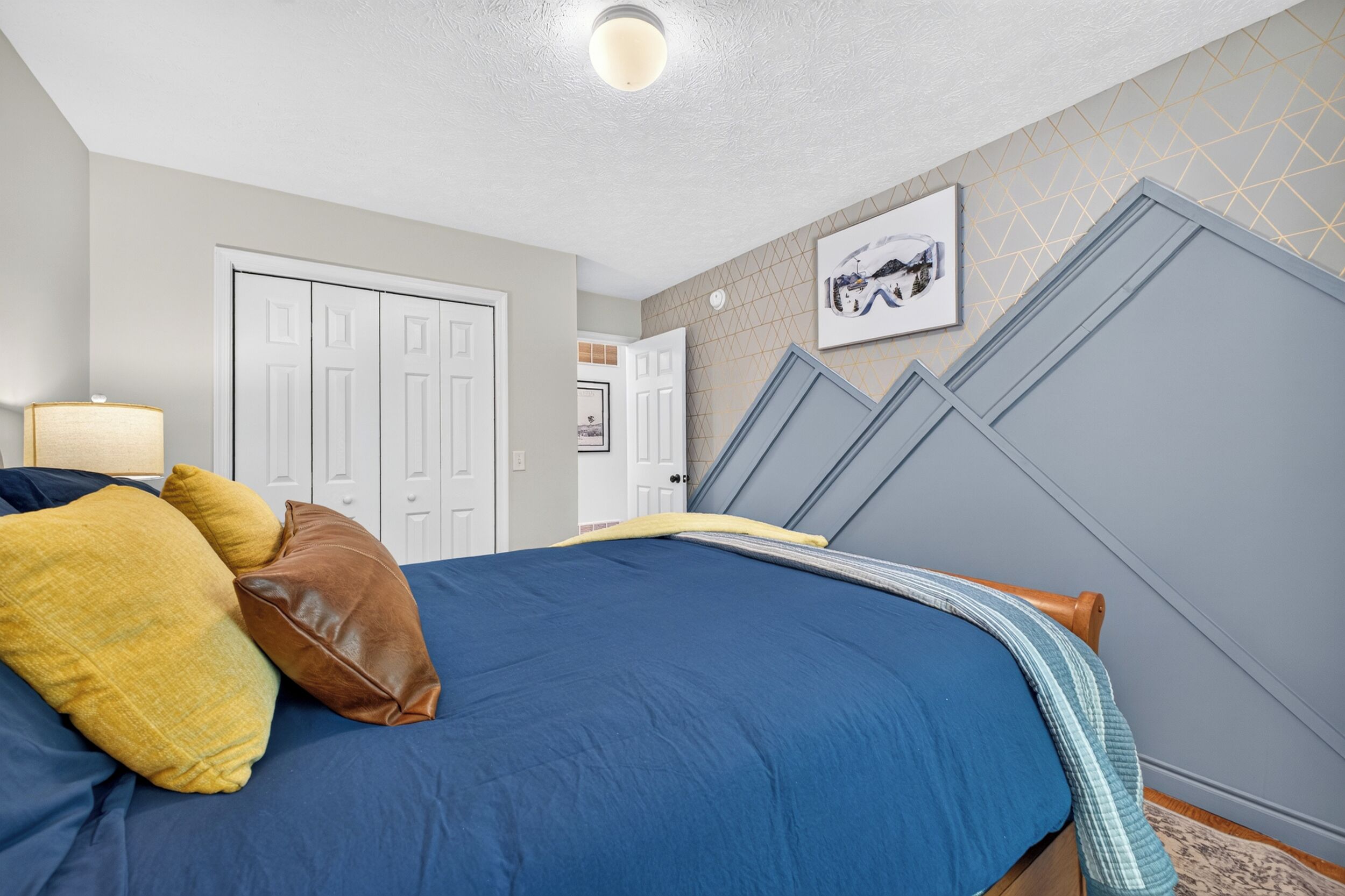 Une chambre à coucher avec un lit, des oreillers, une lampe, une porte, un mur orné d’un motif géométrique et un tableau encadré.