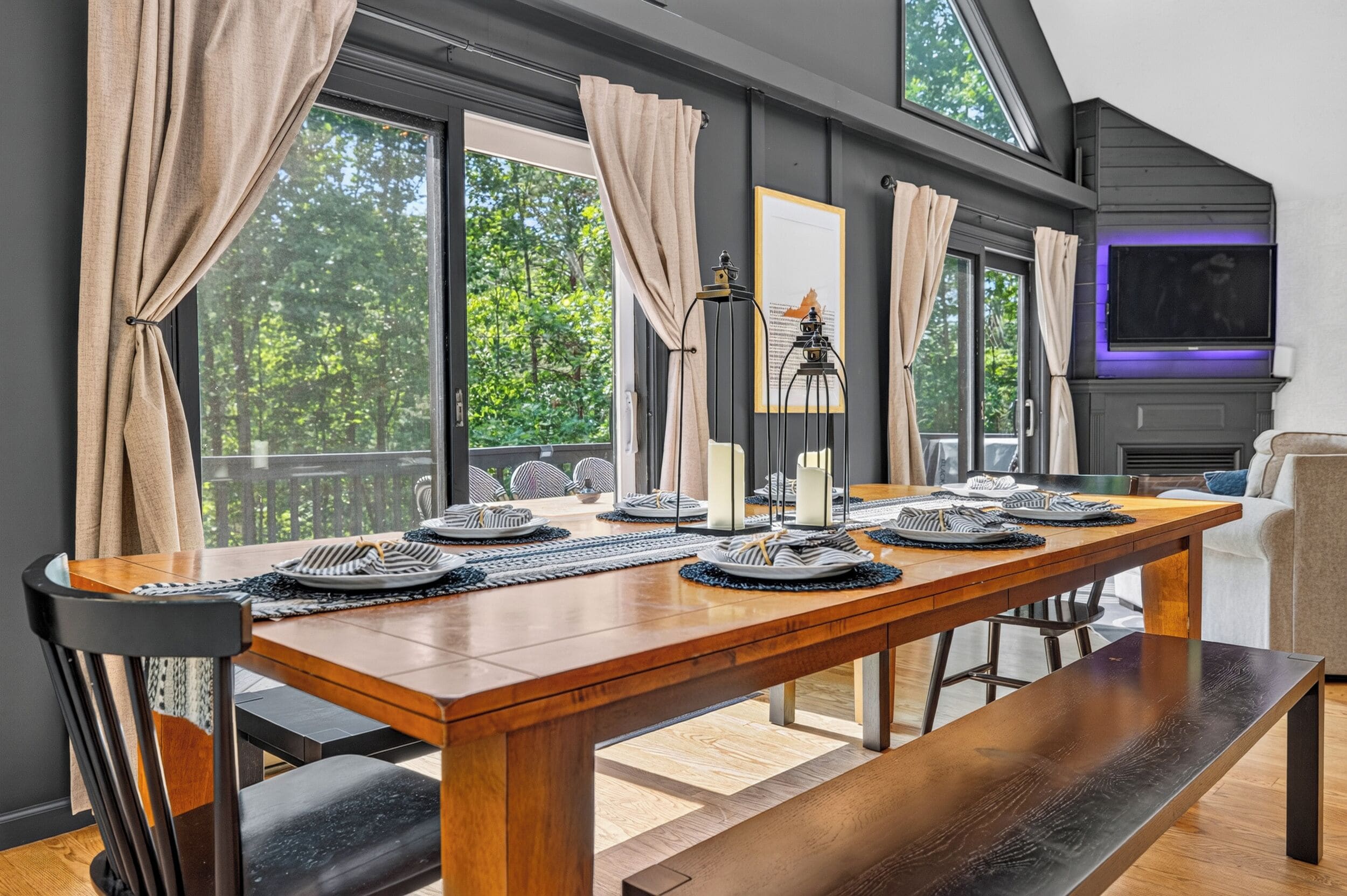 A dining area with a long wooden table set for a meal, large windows with curtains, and a view of trees outside.