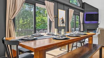 A dining area with a long wooden table set for a meal, large windows with curtains, and a view of trees outside.