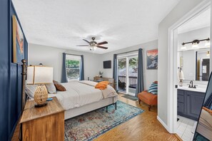 A bedroom with a bed, a nightstand, a lamp, a ceiling fan, a window with curtains, a door leading to a balcony, and a bathroom with a sink and mirror.