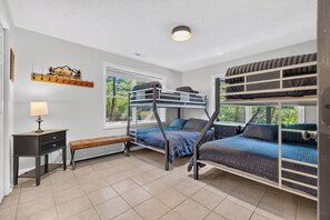 Une chambre avec un lit superposé, un bureau et un banc, un porte-manteau orné d’un tableau représentant un paysage, et une fenêtre donnant sur les arbres.