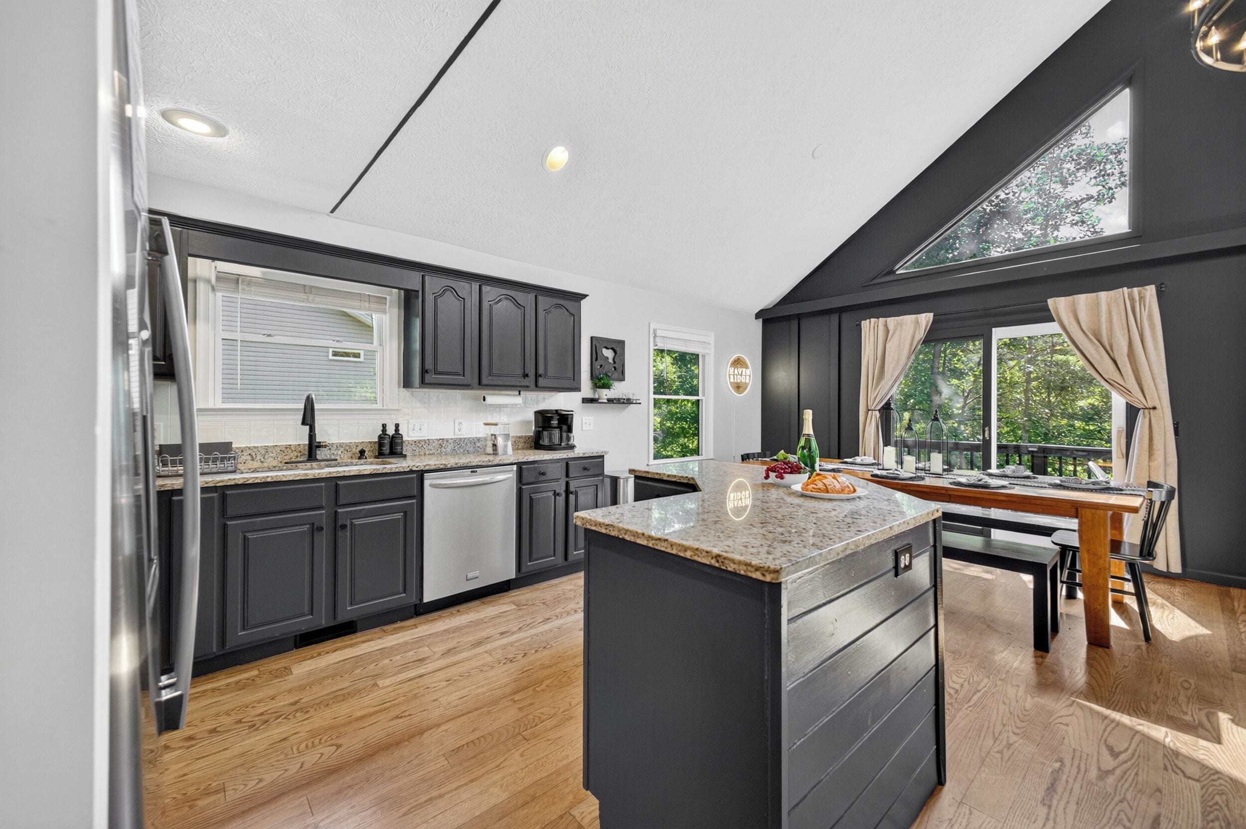 A modern kitchen with dark cabinetry, a central island, and a dining area with a view of greenery.