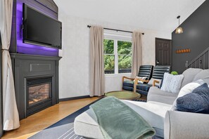 A living room with a fireplace, a television mounted above it, a window with curtains, a sofa, and a staircase.