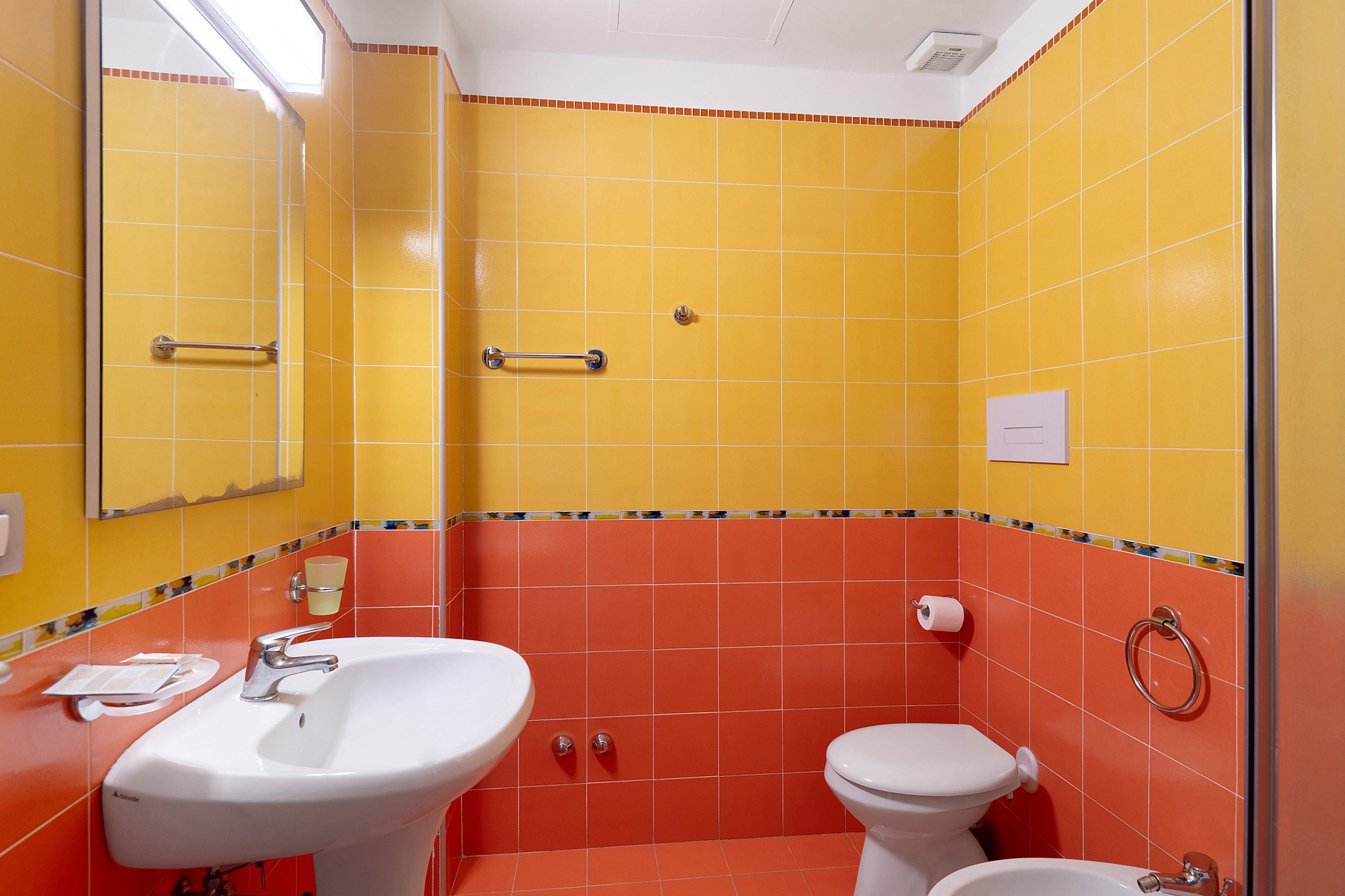 Un baño con azulejos amarillos y anaranjados, un lavamanos blanco, un inodoro y un espejo.
