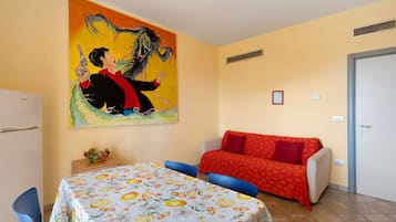 A room with a bed, a sofa, a table with a floral tablecloth, a painting on the wall, and a door.