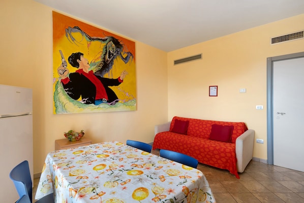 Una habitación con una cama, un sofá, una mesa con mantel floral, un cuadro en la pared y una puerta.