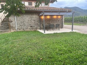 Edifici de pedra amb teulada a dues aigües, zona exterior coberta amb seients i taula, i un jardí amb gespa.