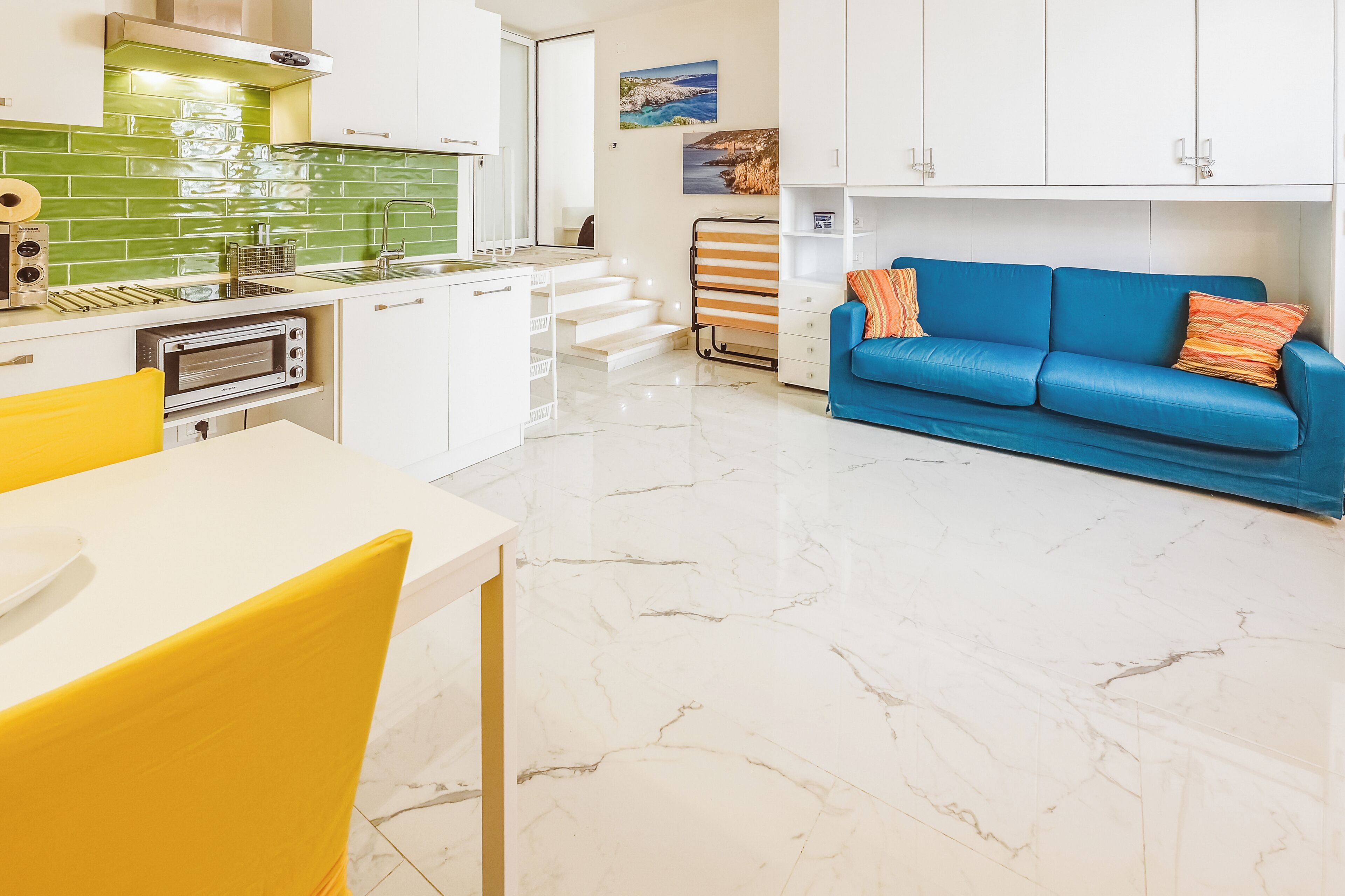 A modern kitchen with a white countertop, a yellow chair, and a blue sofa with orange pillows.