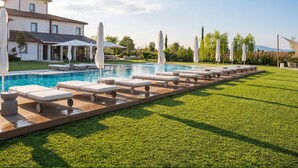 Una piscina con sillones de descanso y sombrillas, una casa y un césped cuidado.
