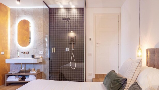 Un dormitorio moderno con una cama grande, una mesita de noche, un espejo, zona de ducha y una puerta.