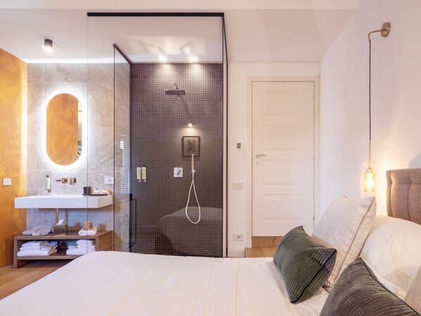 Un dormitorio moderno con una cama grande, una mesita de noche, un espejo, zona de ducha y una puerta.