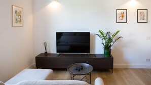 Una sala de estar moderna con un televisor de pantalla plana sobre un mueble de madera oscura, una pequeña mesa redonda con una tetera, dos sillones acolchados, una planta en maceta y obras de arte enmarcadas en las paredes.