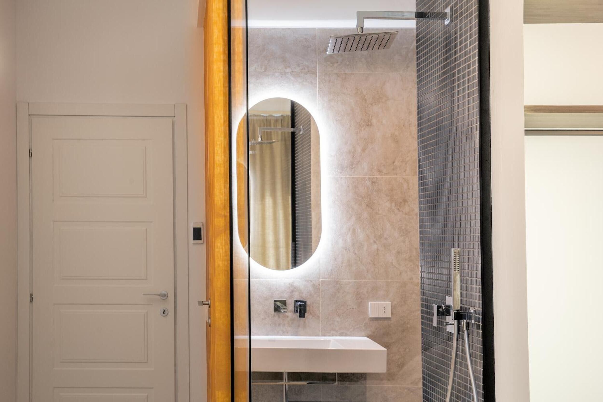 A bathroom with a white door, a mirror with a light, a shower, and a sink.