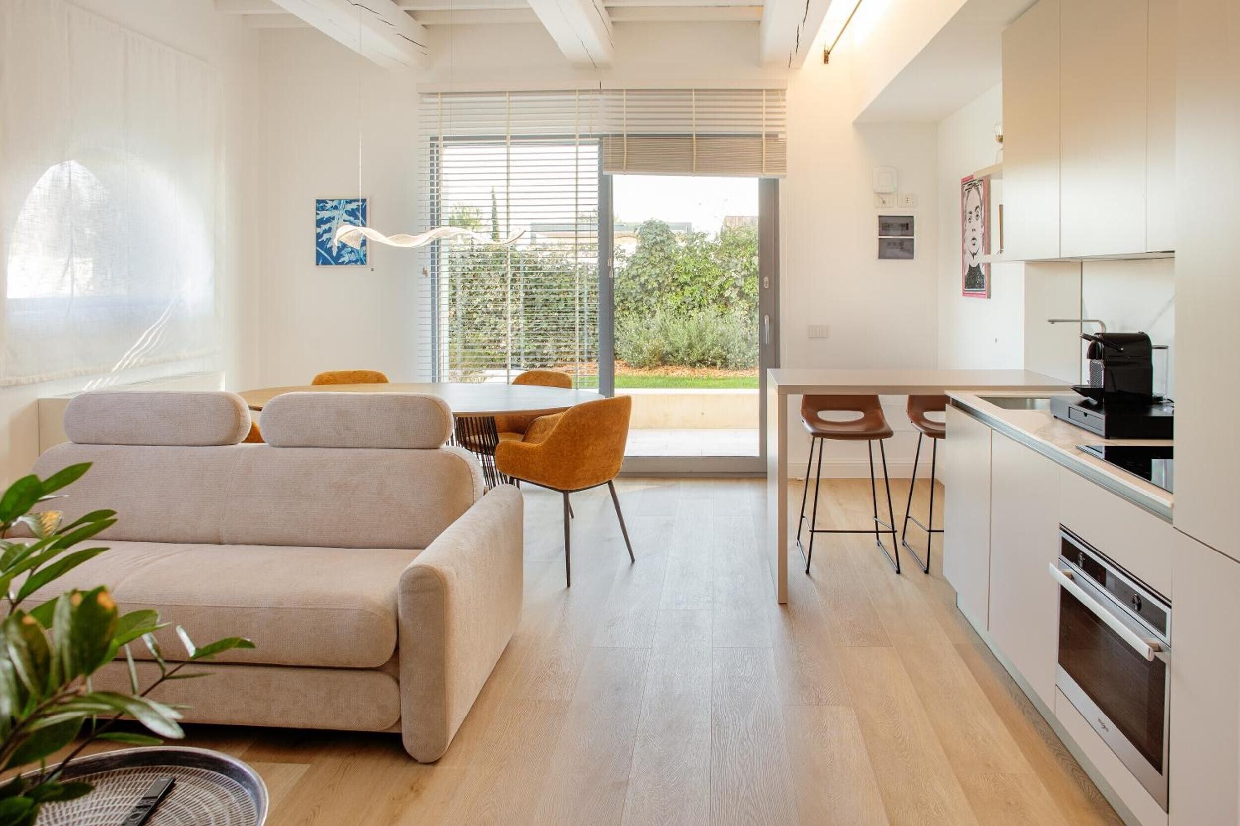 A modern living room with a sofa, a dining table, and chairs. There is a kitchen area with a stove and a refrigerator.