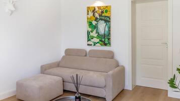 Ein sofa in neutraler Farbgebung mit passendem Hocker, ein abstraktes Wandbild und eine schlicht gestaltete Tür.
