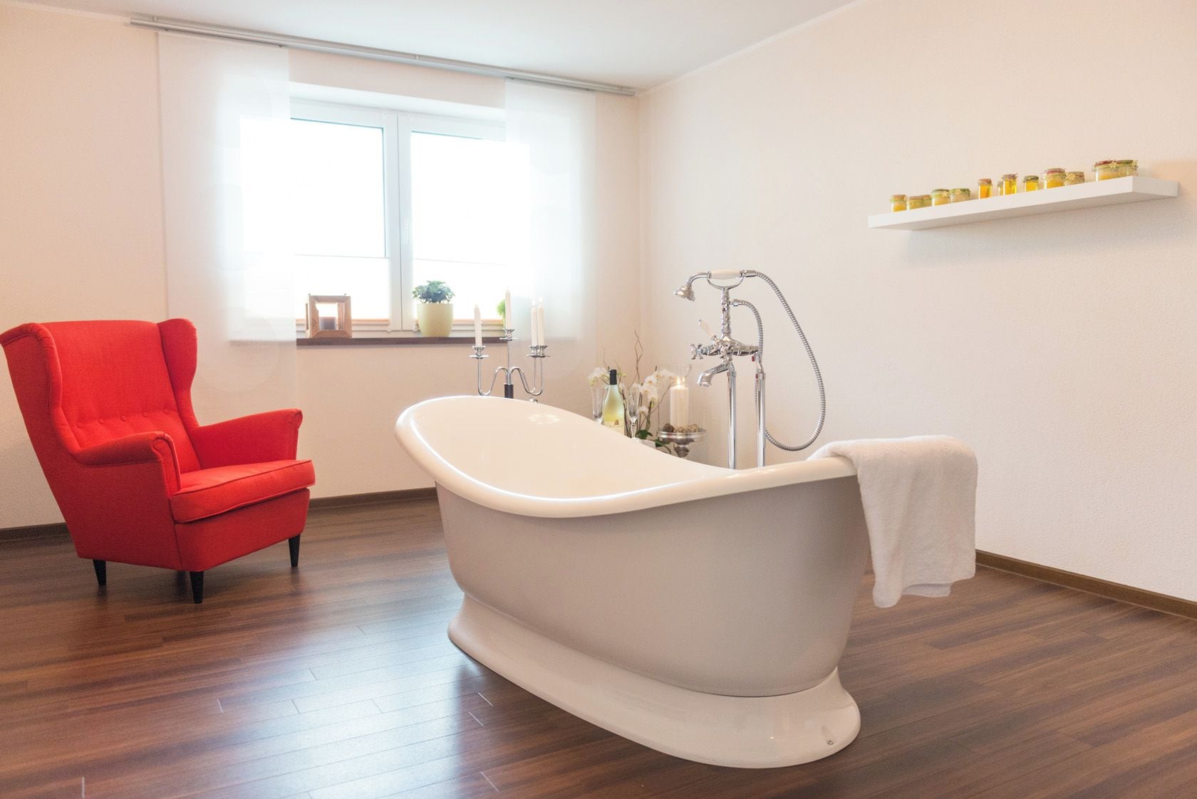 Ein Badezimmer mit einer freistehenden Badewanne, einem roten Sessel, Holzboden, einem Fenster und einem Regal mit Gläsern.