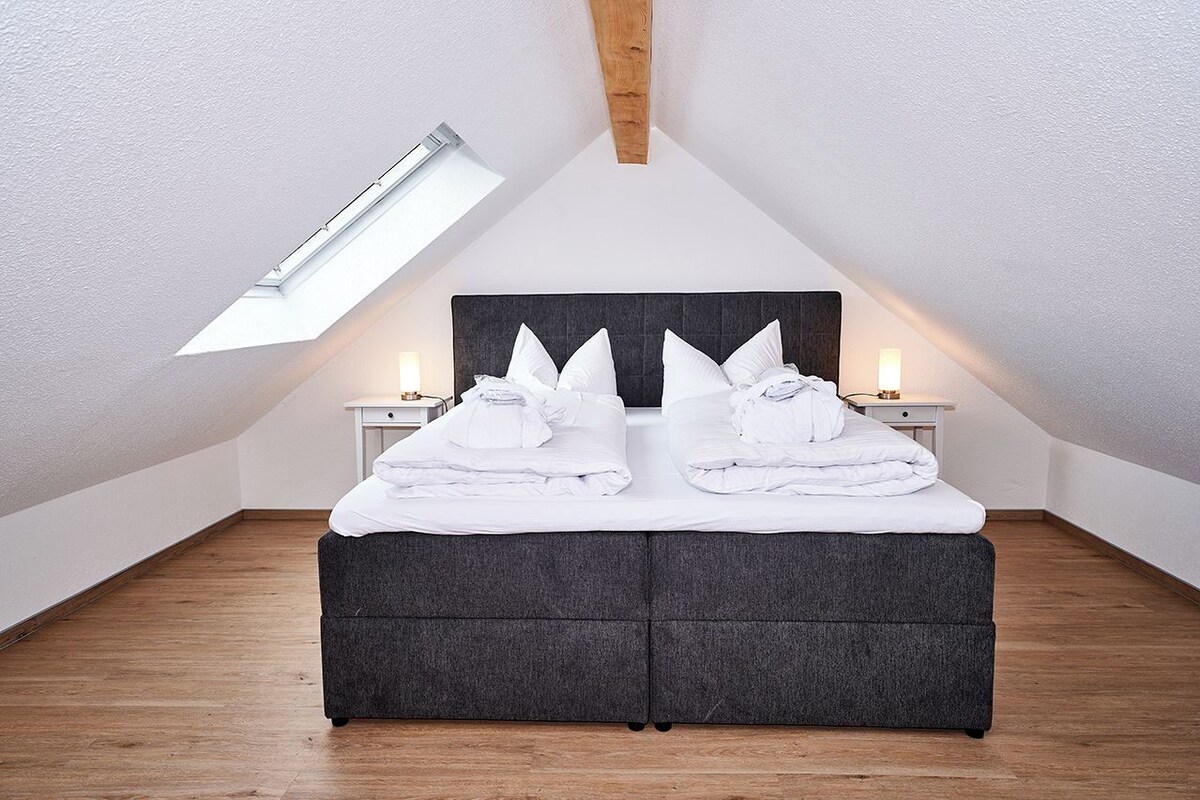 A bedroom with a sloped ceiling, a skylight, a bed with pillows, and bedside lamps.