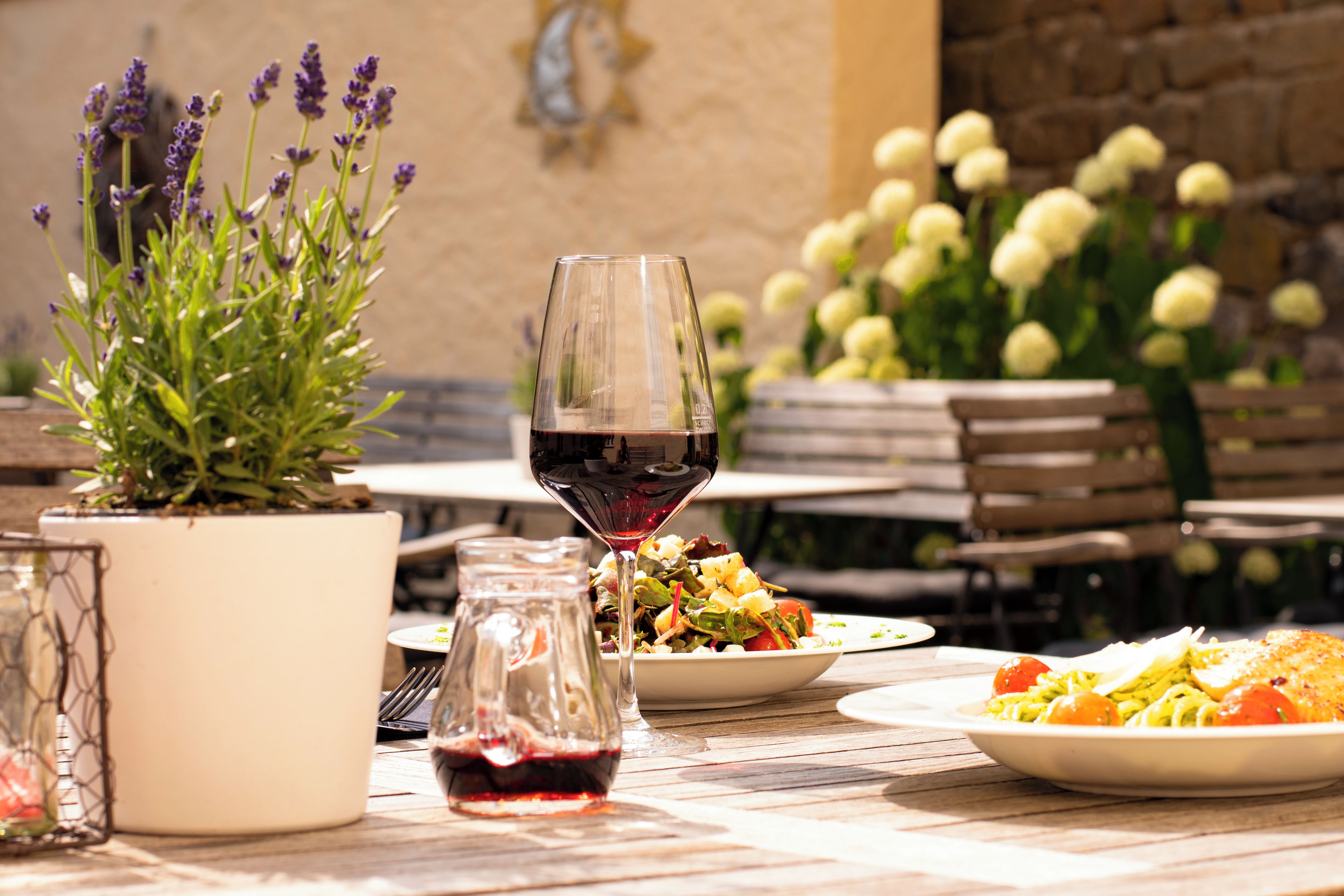 Ein Essbereich im Freien mit Tisch, gedeckten Tellern, einem Glas Rotwein und einem Blumentopf mit violetten Blüten.