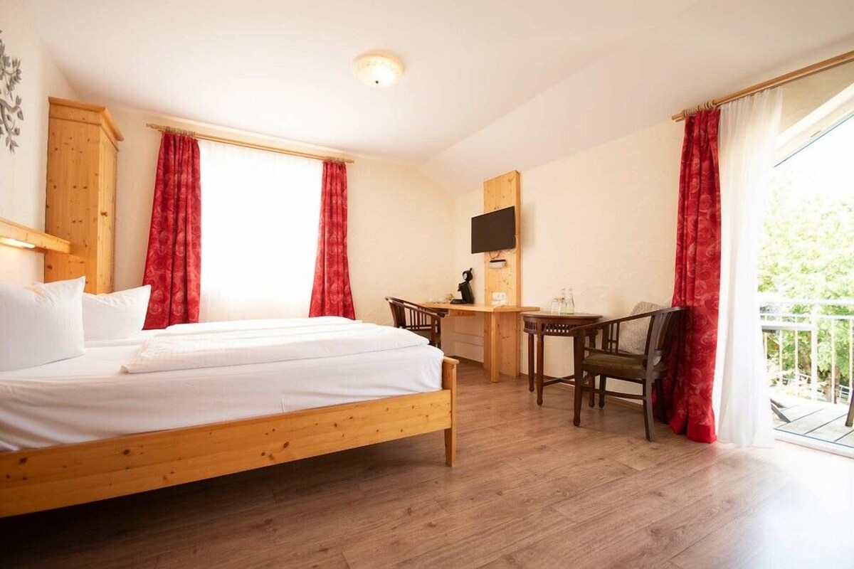 A bedroom with a wooden bed, a desk, and a chair, a television mounted on the wall, and a balcony with red curtains.