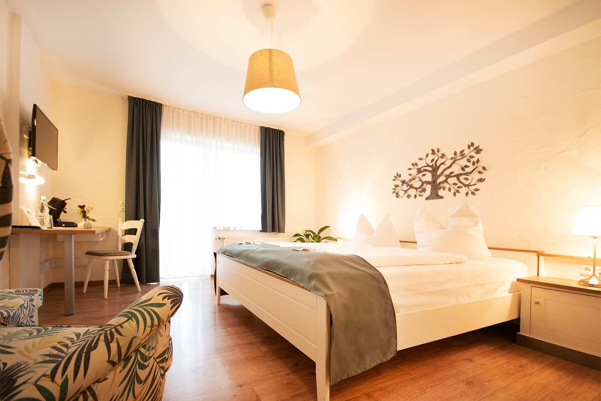 A hotel room with a bed, bedside table, lamp, desk, chair, and a decorative tree wall art piece.