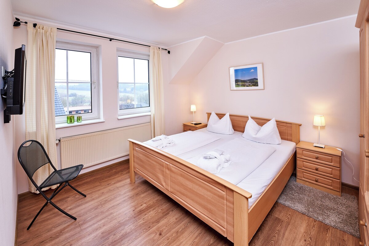 A hotel room with a wooden bed, two bedside tables, a chair, a radiator, and a TV mounted on the wall.