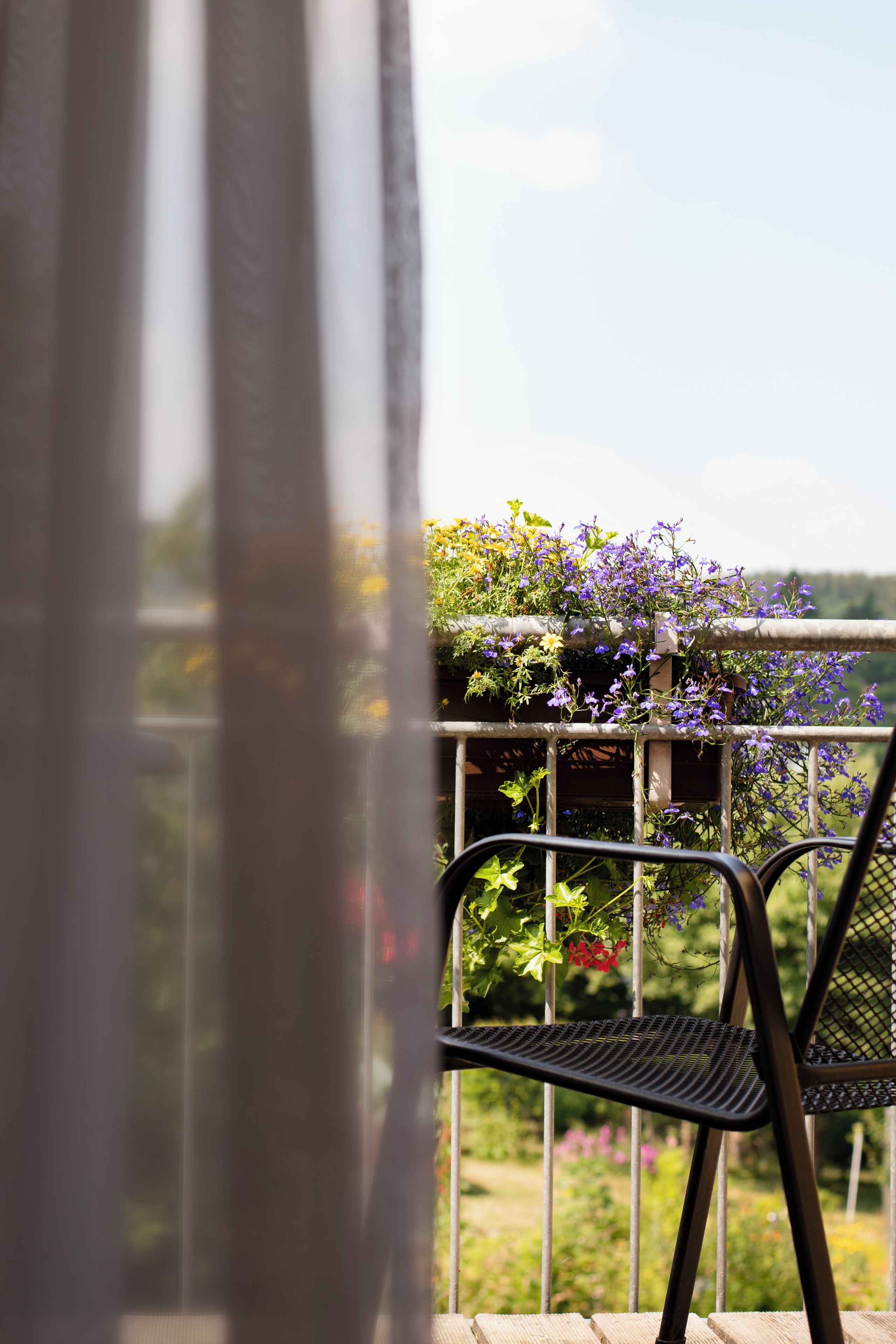 Un balcón con una silla de metal, una jardinera con flores moradas y vista a zonas verdes.