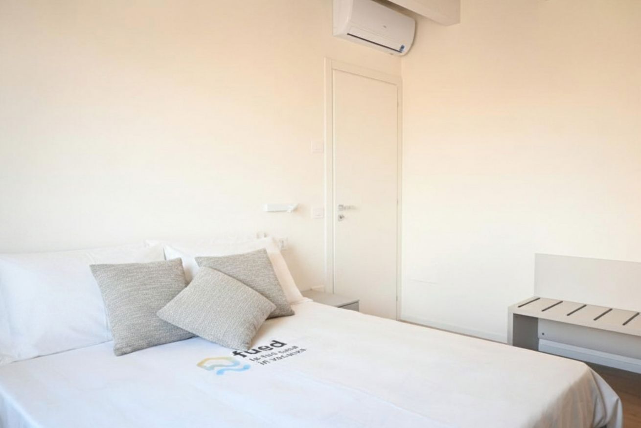 Una habitación de hotel con cama, almohadas, mesita de noche y aire acondicionado instalado en la pared.
