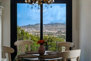 Une table avec un vase et des chaises, placée devant une fenêtre donnant sur un paysage urbain et des collines.