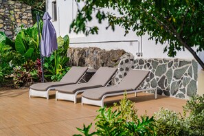 Un espace patio aménagé avec trois chaises longues, un mur en pierre et une végétation luxuriante.