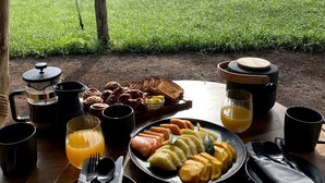 A round wooden table set outdoors with a meal consisting of toast, sausages, and sliced fruit, accompanied by a coffee pot, a thermos, and a glass of orange juice.