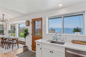 A modern kitchen with a large window, white cabinetry, and a view of the sea. - Perch - 2BR Waterfront Bluff | Sunset Views | Free Parking (Coupeville)