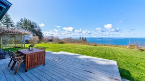 A deck with a table and chairs, overlooking a well-maintained lawn and a body of water. - Perch - 2BR Waterfront Bluff | Sunset Views | Free Parking (Coupeville)