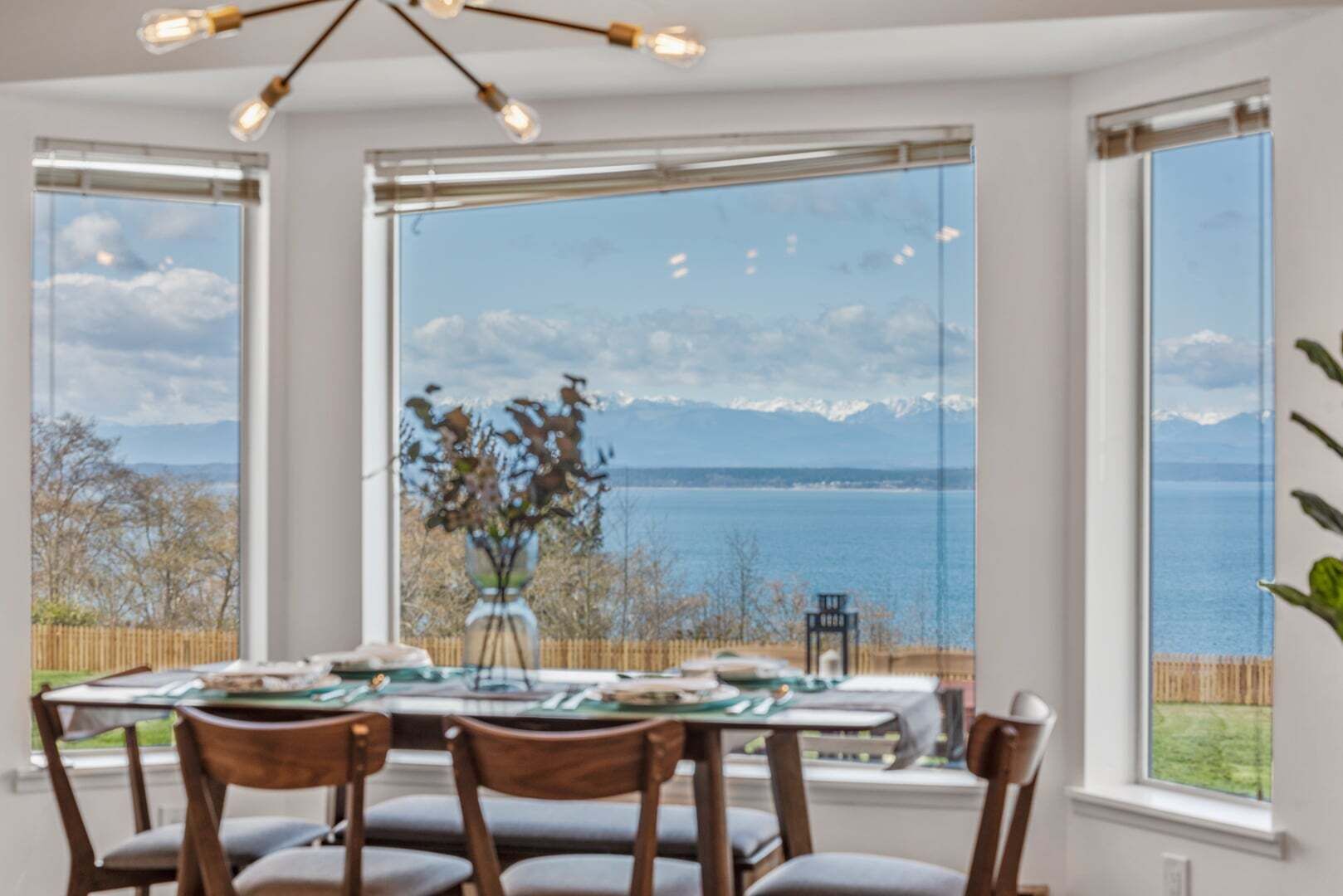 Spisested med et bord dækket til fire personer, store vinduer med udsigt til vand og bjerge, og en hængende lysekrone.