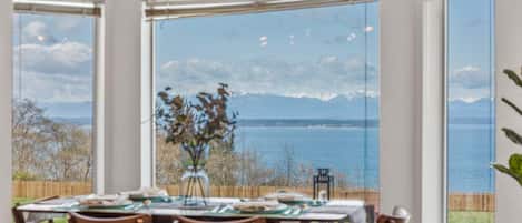 Un comedor con una mesa dispuesta para cuatro, amplias ventanas con vistas a un cuerpo de agua y montañas, y una lámpara colgante.