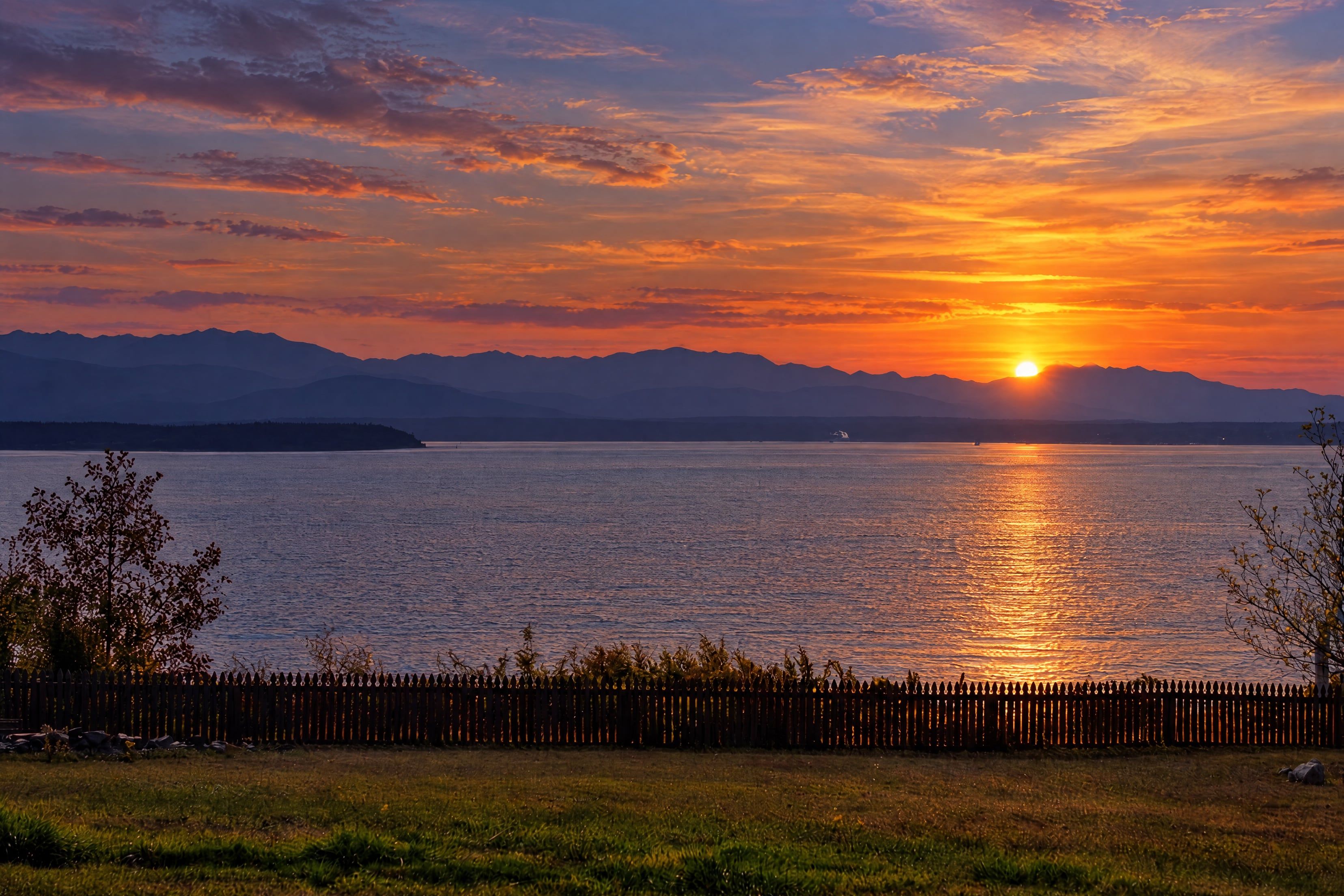 A sunset over a calm lake with mountains in the distance and a wooden fence in the foreground.