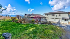 A two-story house with a garage, a well-maintained lawn, and a clear view of the mountains in the background. - Perch - 2BR Waterfront Bluff | Sunset Views | Free Parking (Coupeville)