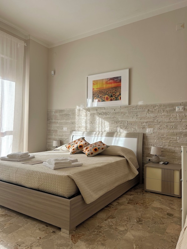A bedroom with a bed, a nightstand, a lamp, a framed picture on the wall, and a window with curtains.