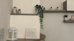 Una habitación con chimenea, un reloj, estanterías con plantas, una cita enmarcada y un farol blanco.