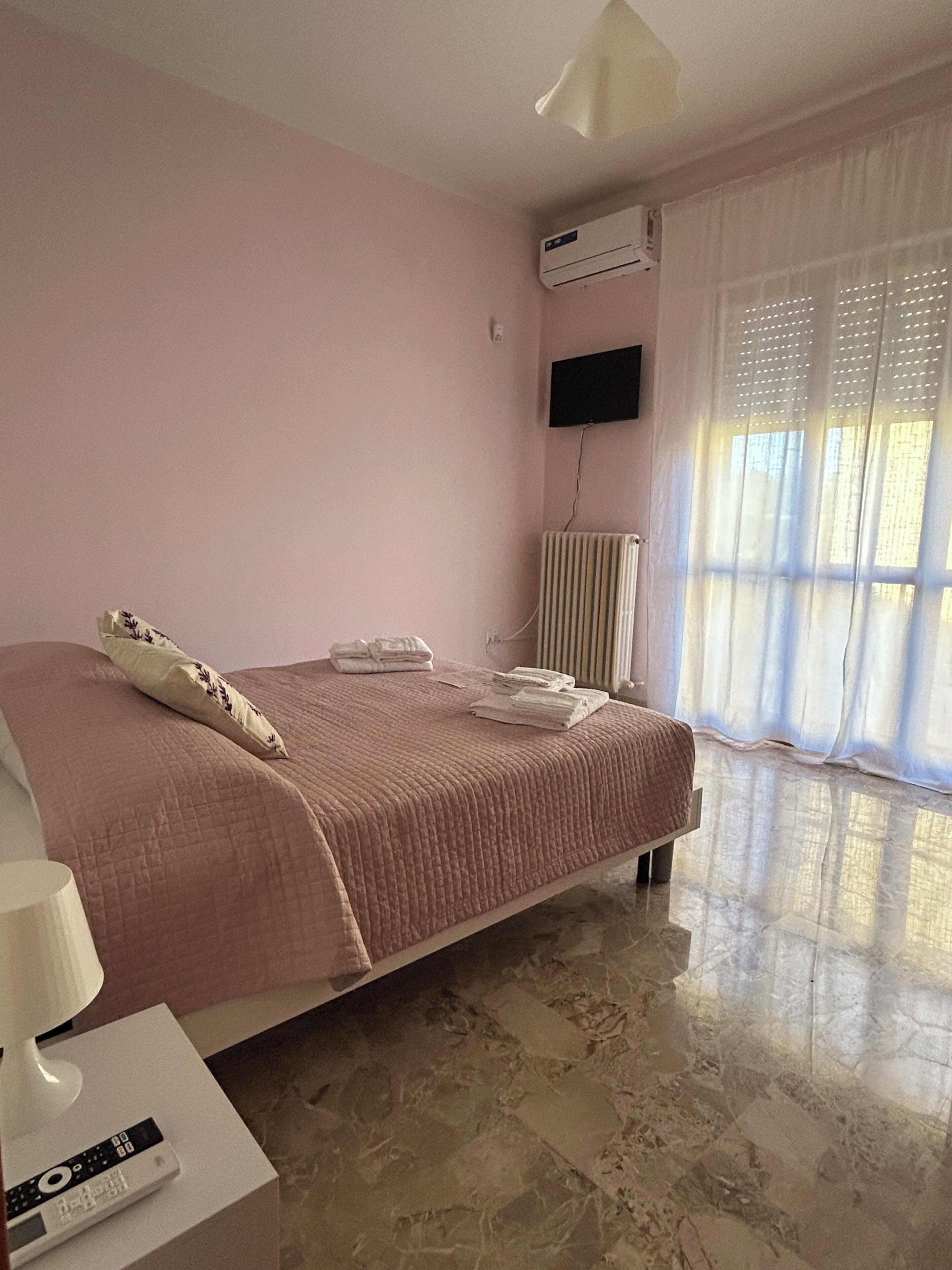 A bedroom with a bed, a flat-screen TV, an air conditioner, a radiator, and a window with sheer curtains.