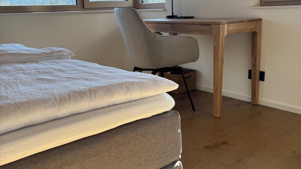 A bedroom with a bed, a desk, and a chair. There are large windows with a mountain view. - Mountain Ranges Around Lake Chiemsee (Übersee)