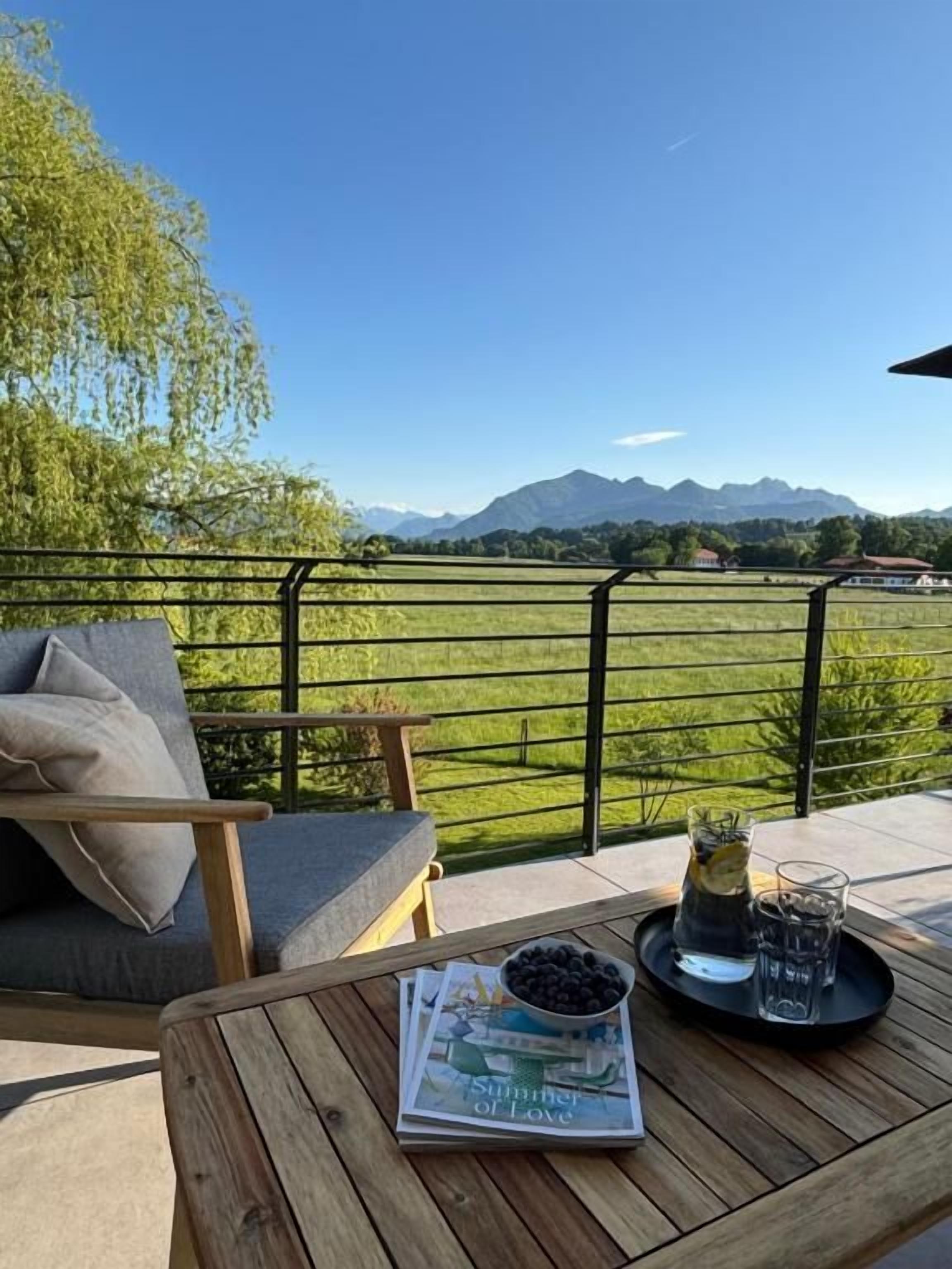 A wooden table with a magazine, a bowl of blueberries, and a glass of water on a balcony with a view of mountains and a tree.