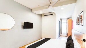 Habitación de hotel con cama, ventilador de techo, equipo de aire acondicionado, televisor de pantalla plana montado en la pared y una puerta que da a otra habitación.