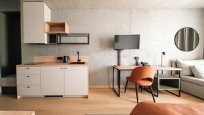 Un espace de vie moderne et compact, comprenant un coin cuisine, un téléviseur à écran plat, un miroir rond et un espace détente avec un canapé et un fauteuil.