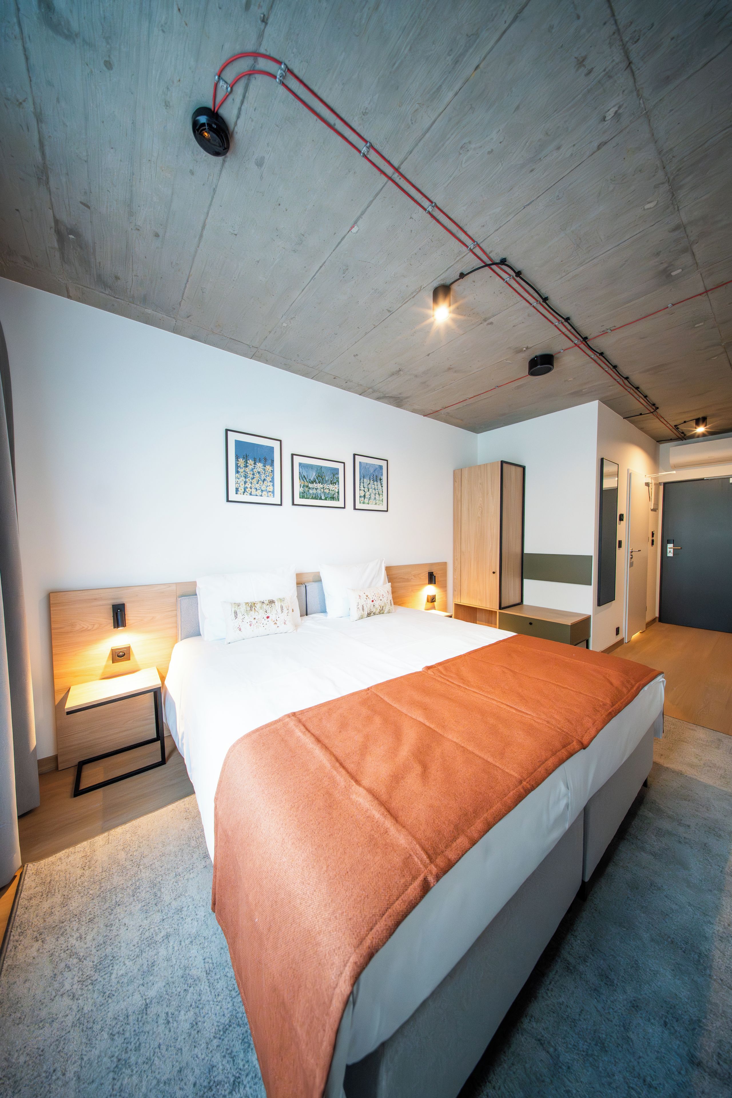 Una habitación de hotel moderna con una cama grande, un armario de madera, una mesita de noche y una lámpara de pared.