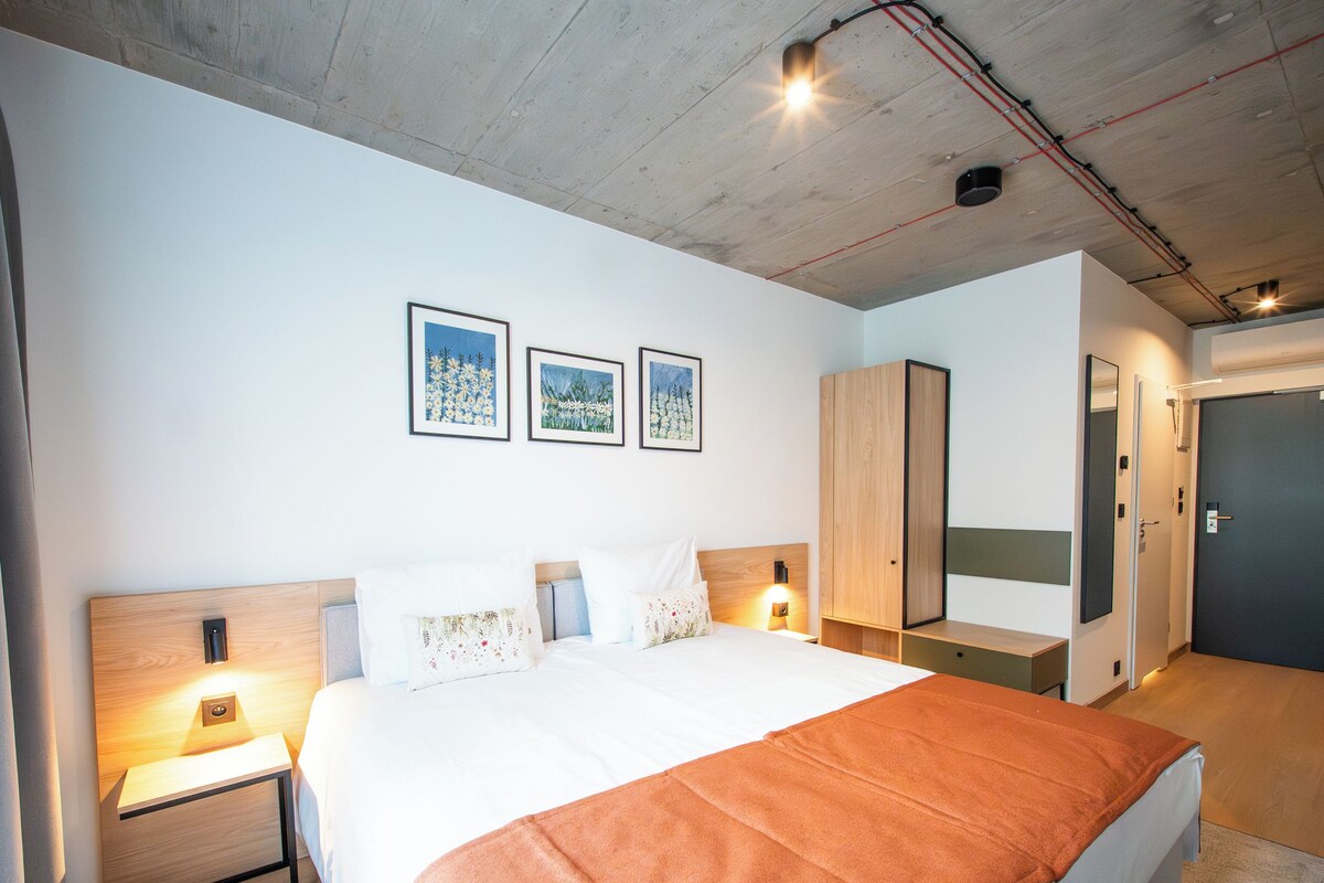 Quarto de hotel moderno com uma cama grande, um armário de madeira, uma mesa de cabeceira e um aplique na parede.