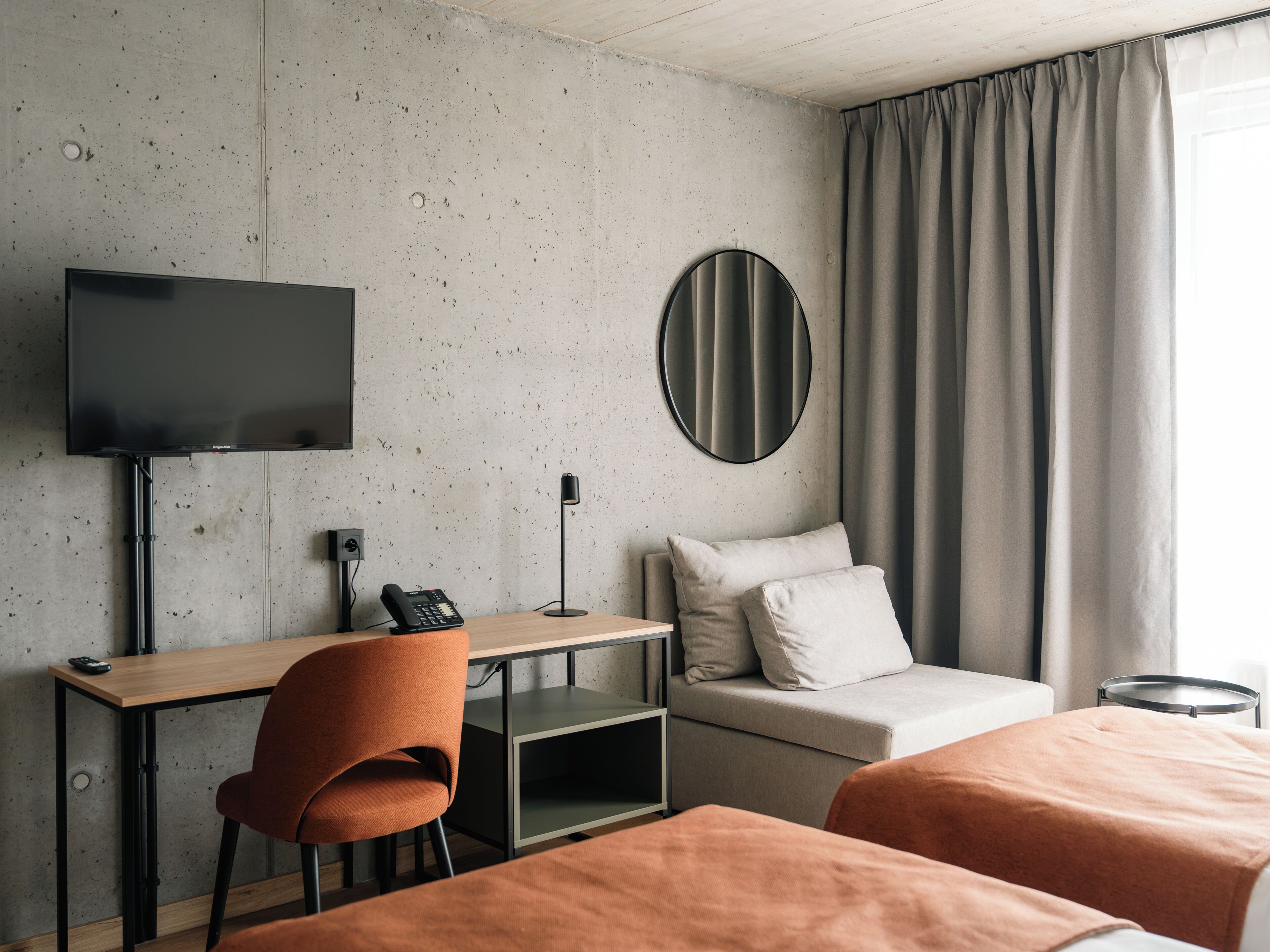 A hotel room with a bed, a desk with a chair, a television, a round mirror, and a window with curtains.