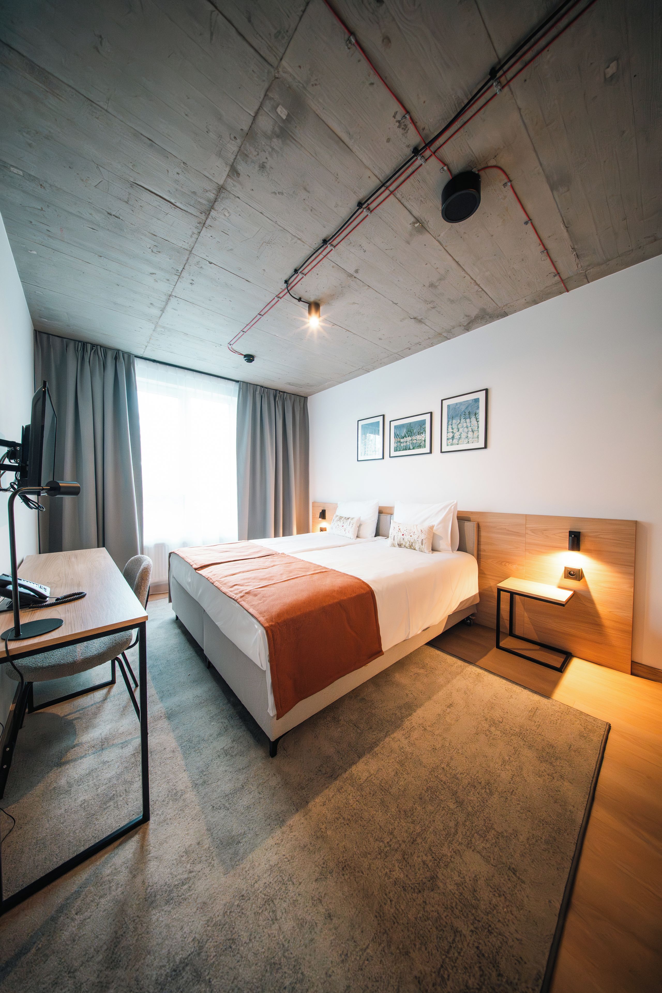 A modern hotel room with a large bed, a desk with a telephone, and a headboard with integrated lighting.
