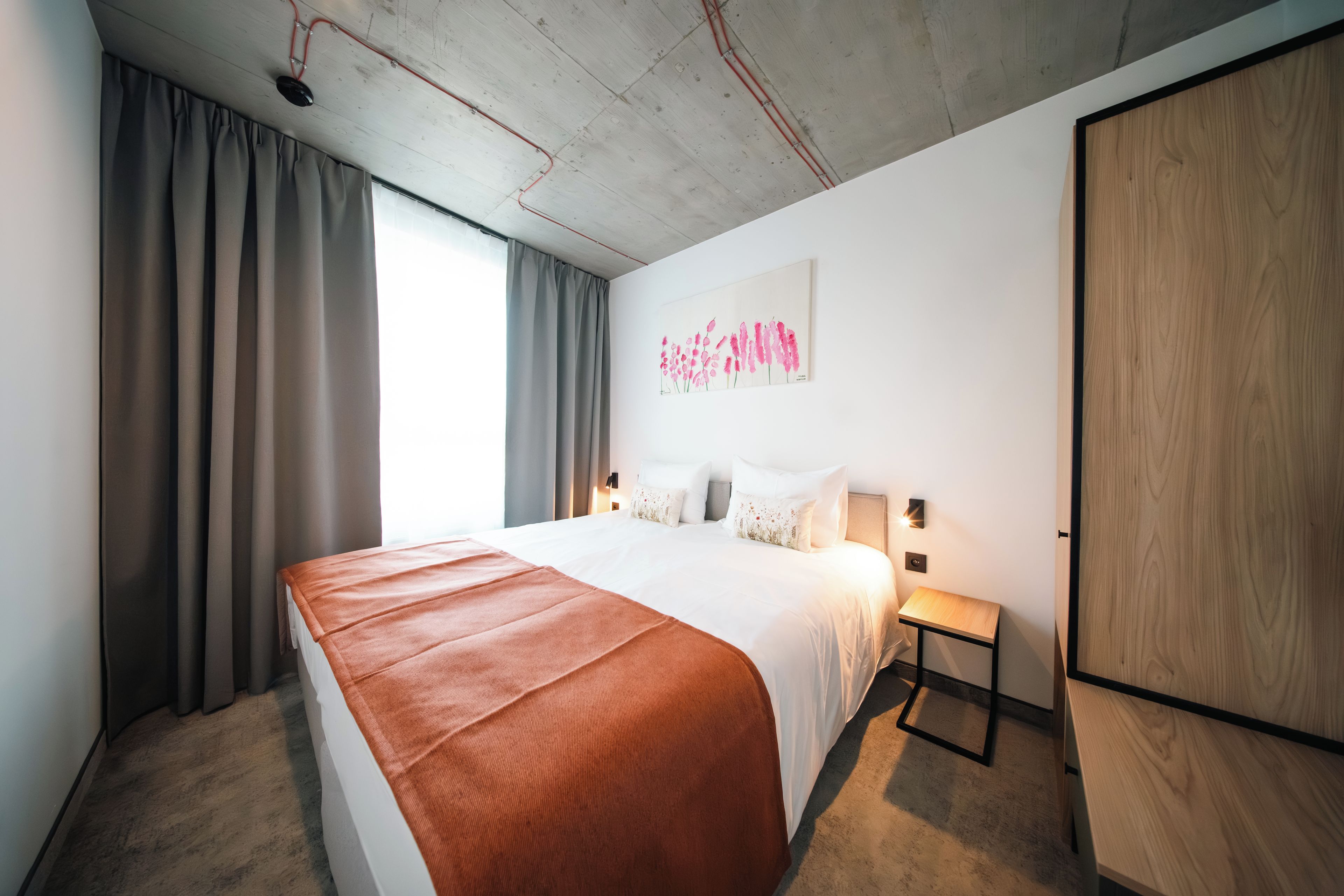 A hotel room with a large bed, a bedside table, a nightstand, a wall-mounted lamp, a painting, and a wardrobe.